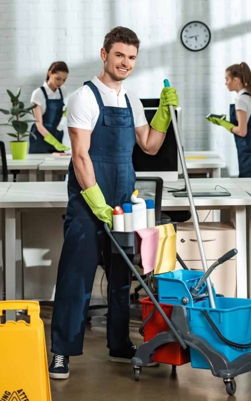 cheerful-cleaner-moving-cart-with-cleaning-supplies-near-multicultural-colleagues.jpg
